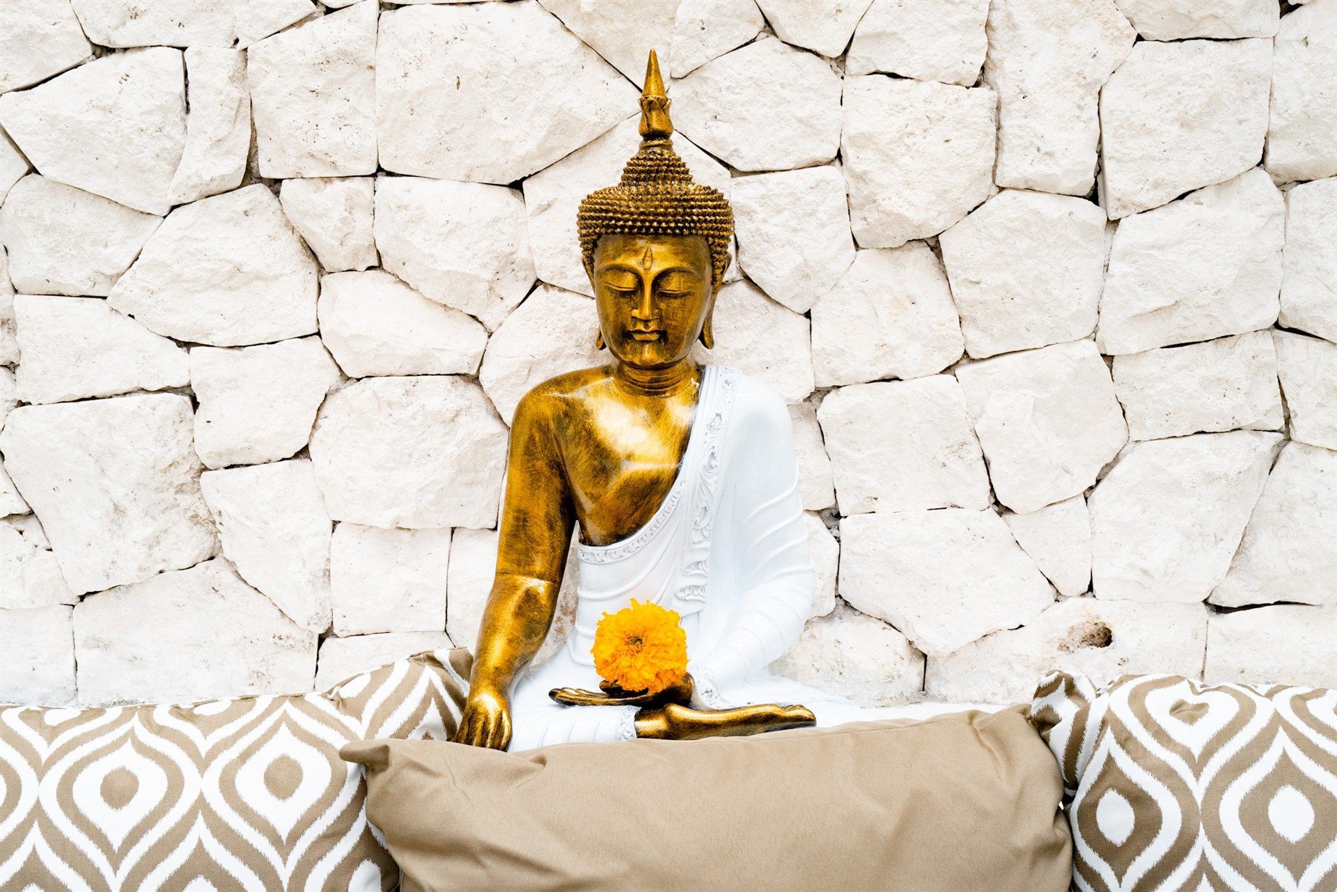 Golden Buddha statue seated with orange flower, in front of a white stone wall.