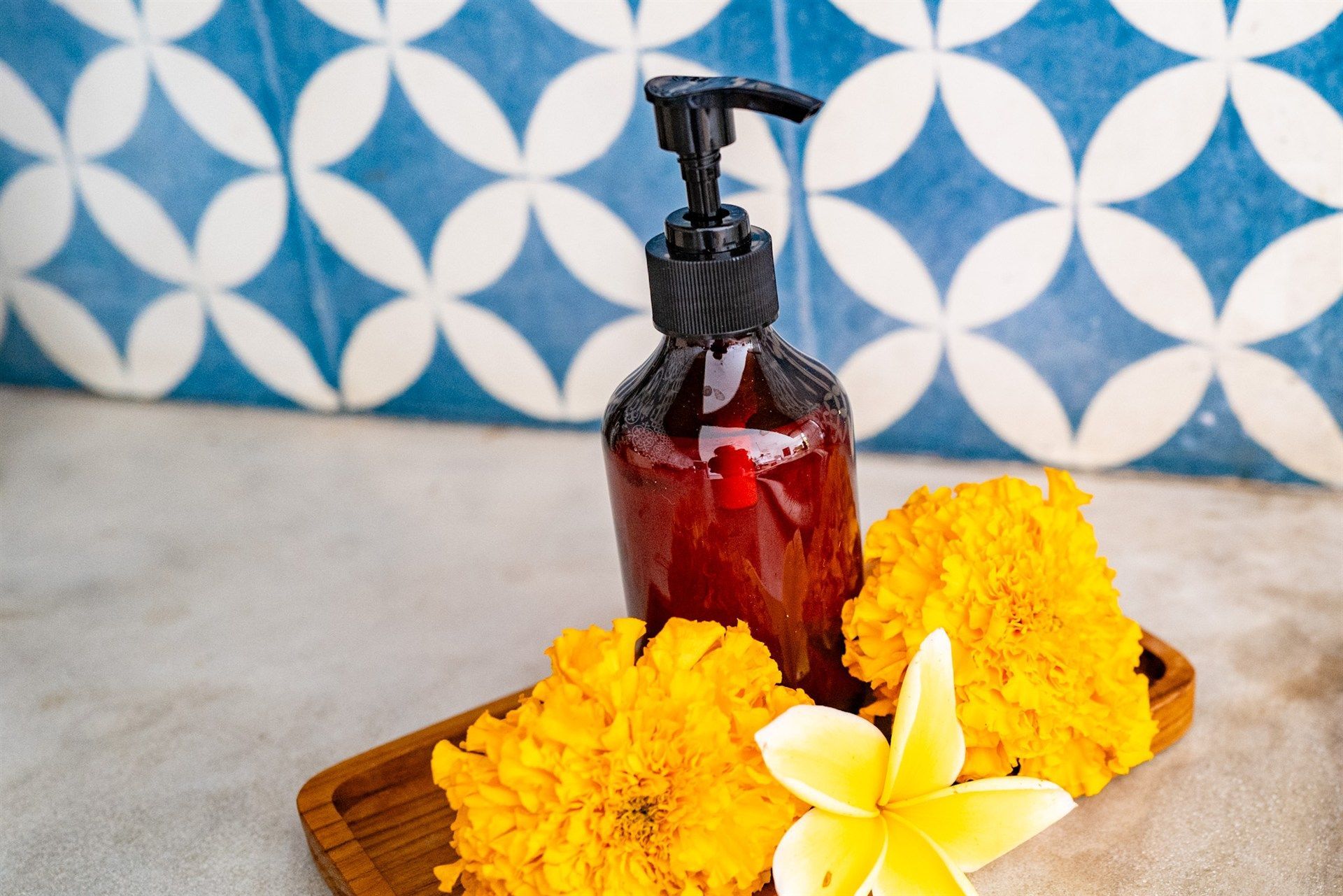 Brown bottle with a black pump, surrounded by yellow flowers, on a wooden tray, against a blue tile backdrop.