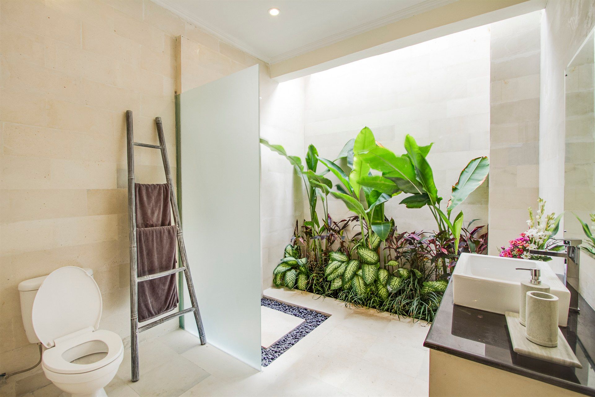 Bathroom with toilet, sink, and an open-air shower area with plants.