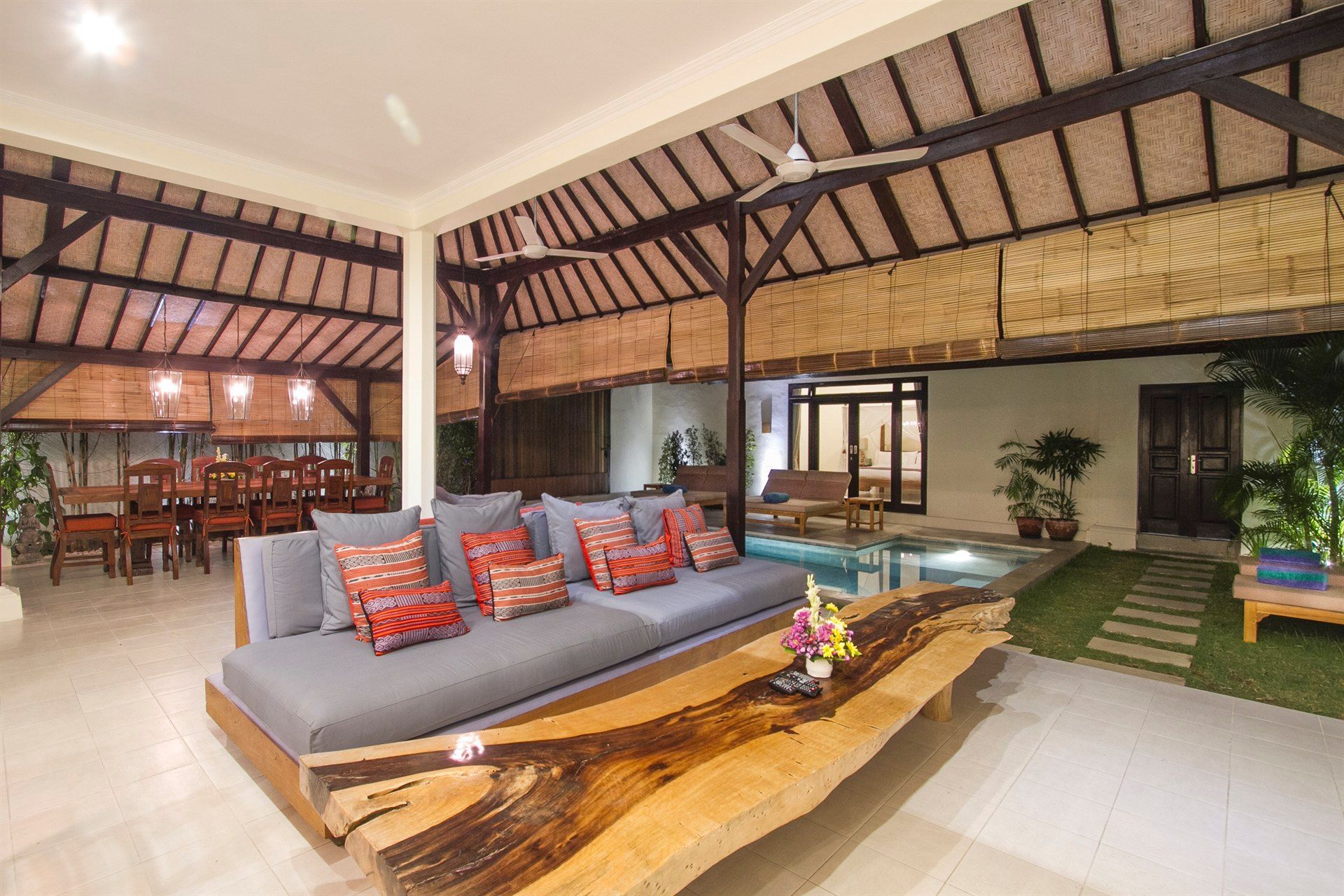 Open-air living area with a long gray couch, wooden table, and view of a pool.