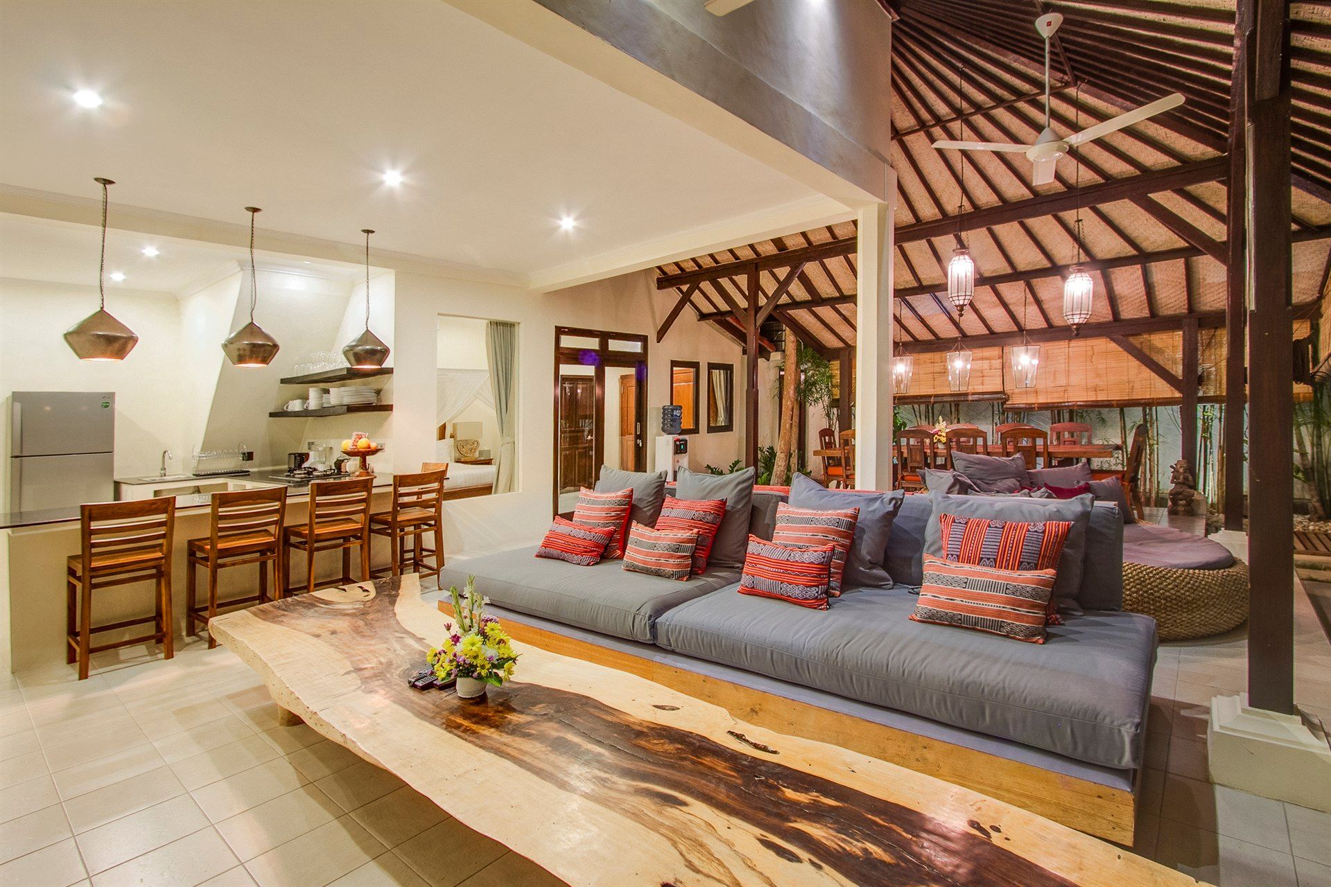 Living area with open kitchen, large wooden table, gray couch with patterned pillows, and exposed wooden ceiling.