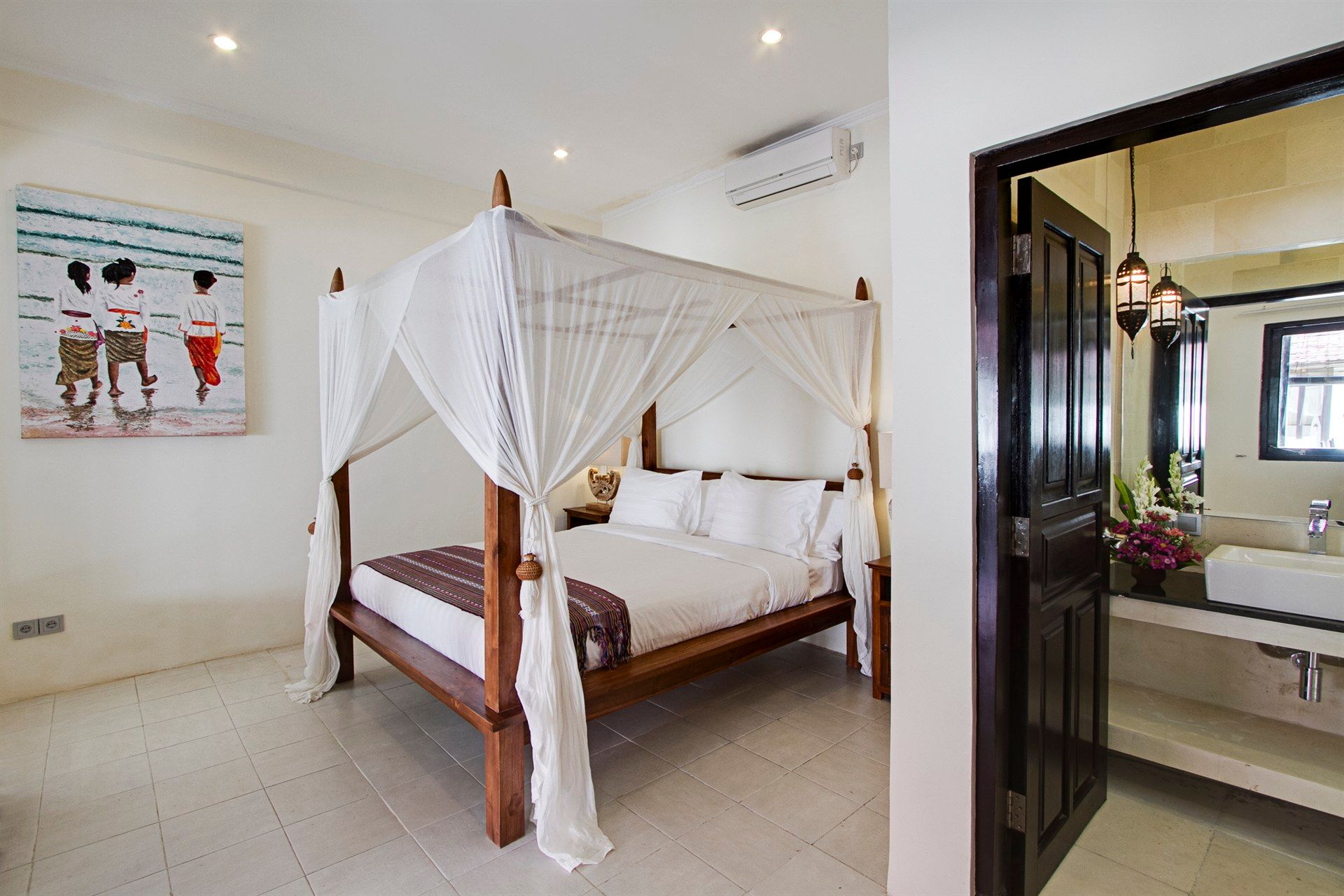 Bedroom with canopy bed, artwork, and bathroom doorway. White walls and wooden furniture.