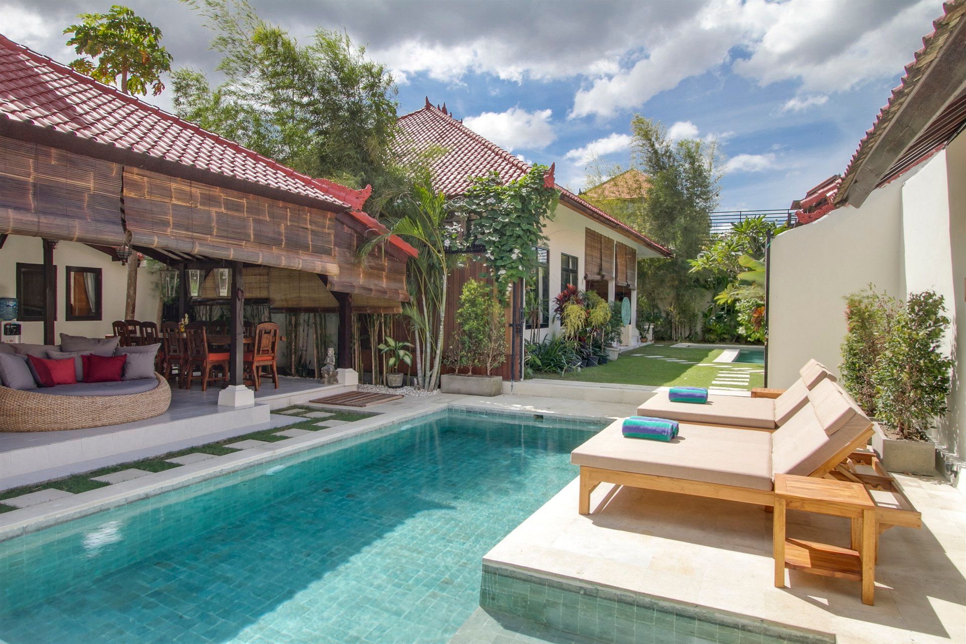 Poolside villa with turquoise water, wooden sunbeds, red-roofed buildings, and lush greenery.