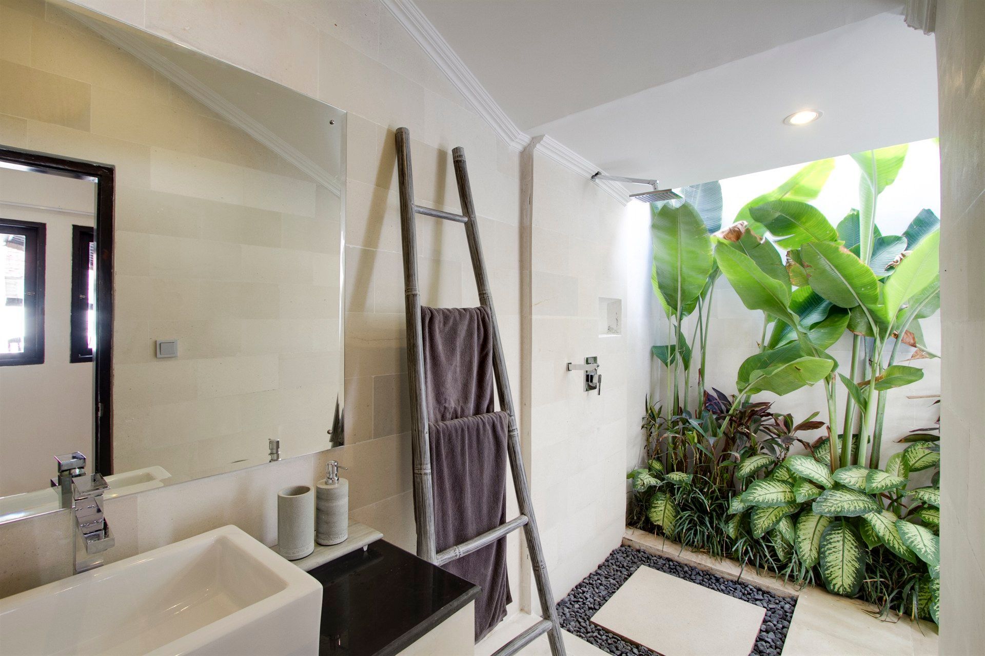 Bathroom with outdoor shower: beige walls, white sink, grey ladder with towels, green plants.