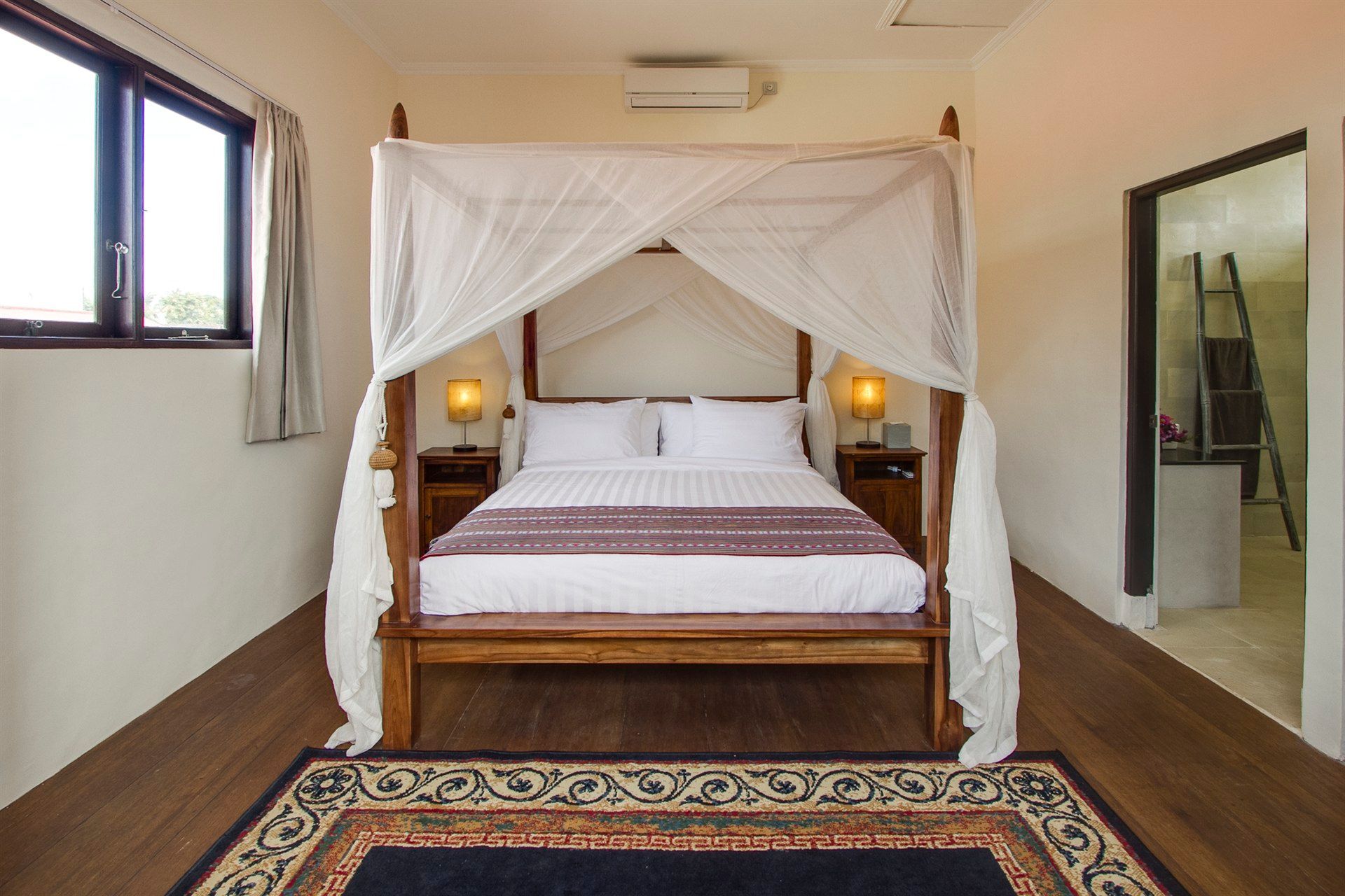 Bedroom with four-poster bed, mosquito net, and rug on a hardwood floor. A window is on the left.