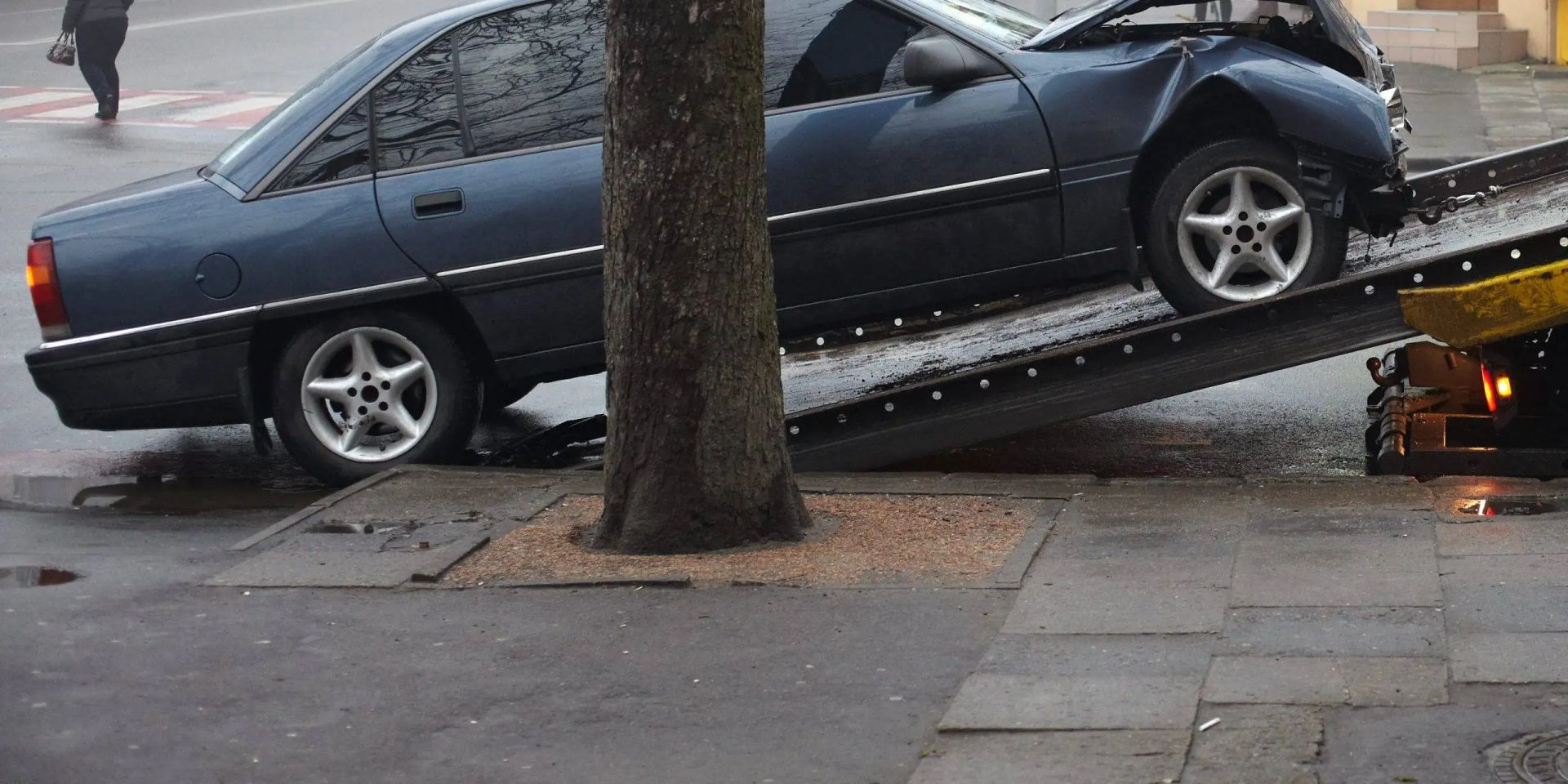 A damaged dark blue car being loaded onto a tow truck, next to a tree on a city street.