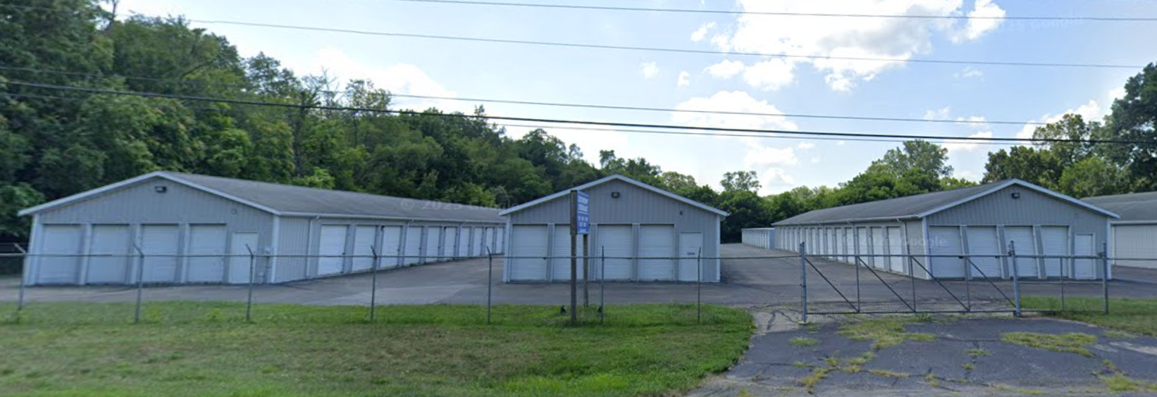 A row of grey self-storage units with white roll-up doors, set against a backdrop of trees under a blue sky.