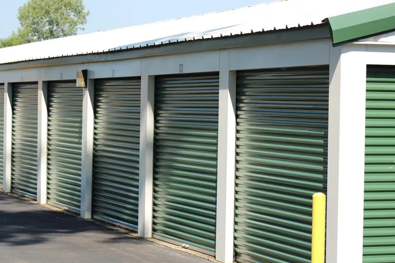 A row of self-storage units with green metal roll-up doors and white framing under a metal roof.