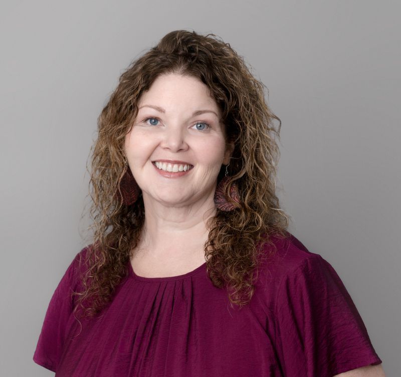 Woman with curly hair smiles, wearing a purple top and earrings, against a gray backdrop.