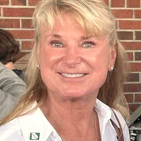 Woman with blonde hair, smiling at camera, wearing white shirt. Brick wall background.