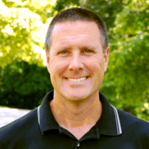 Man smiling, wearing a black polo shirt, with a blurred green background.