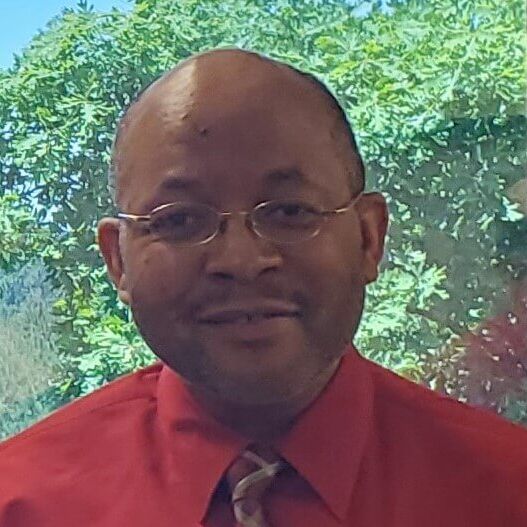Man wearing glasses and a red shirt, smiling, with a nature background.