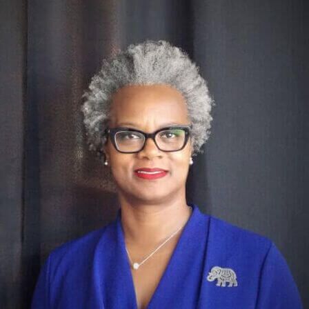 Woman with gray curly hair, wearing glasses and a blue blazer, smiles.