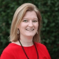 Woman with blonde hair, smiling, wearing red shirt and black necklace, outdoors.