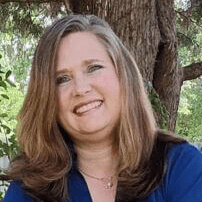 Woman with light brown hair smiles, blue shirt, outdoors near a tree.