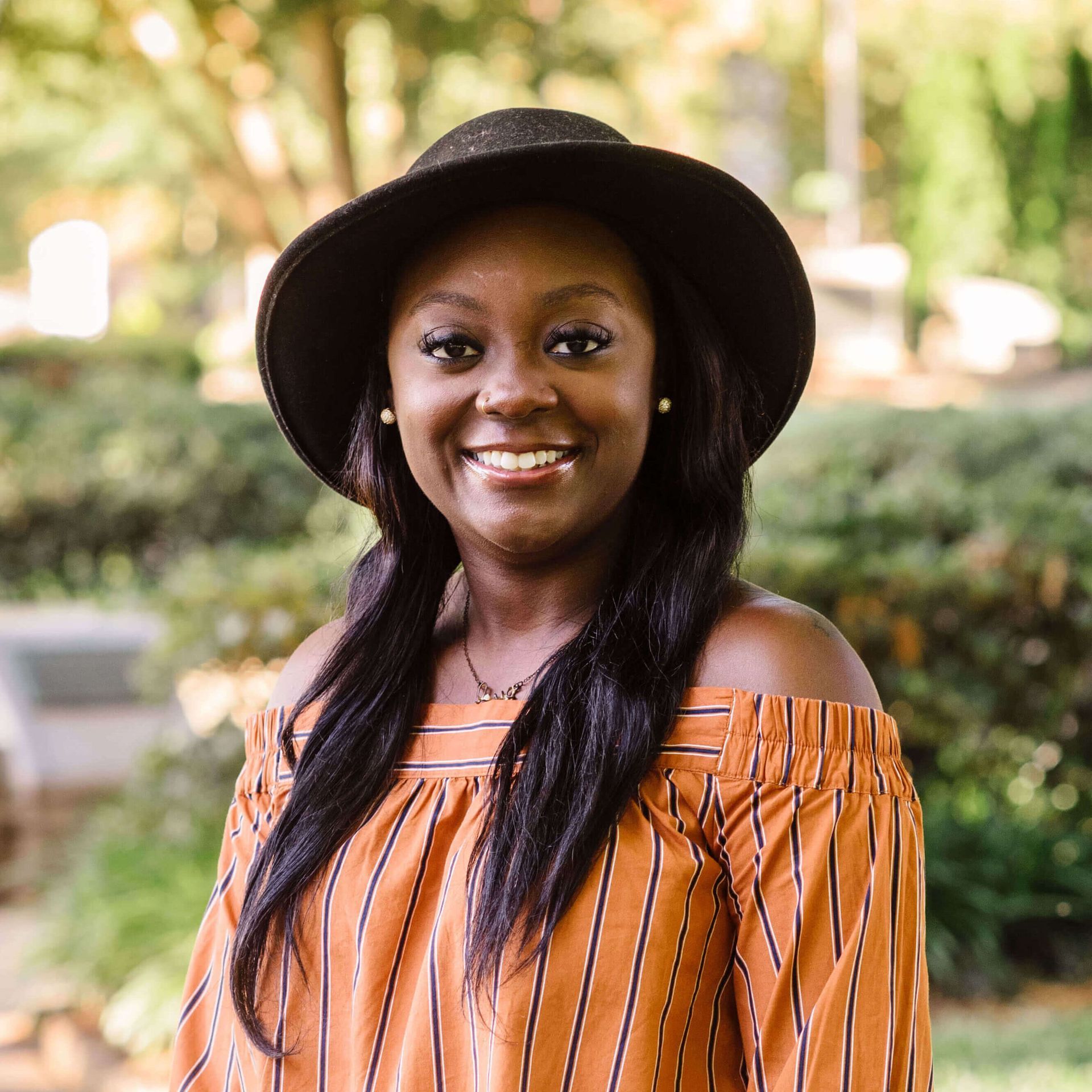 Woman with a black hat smiles outdoors, wearing an orange striped off-shoulder top.