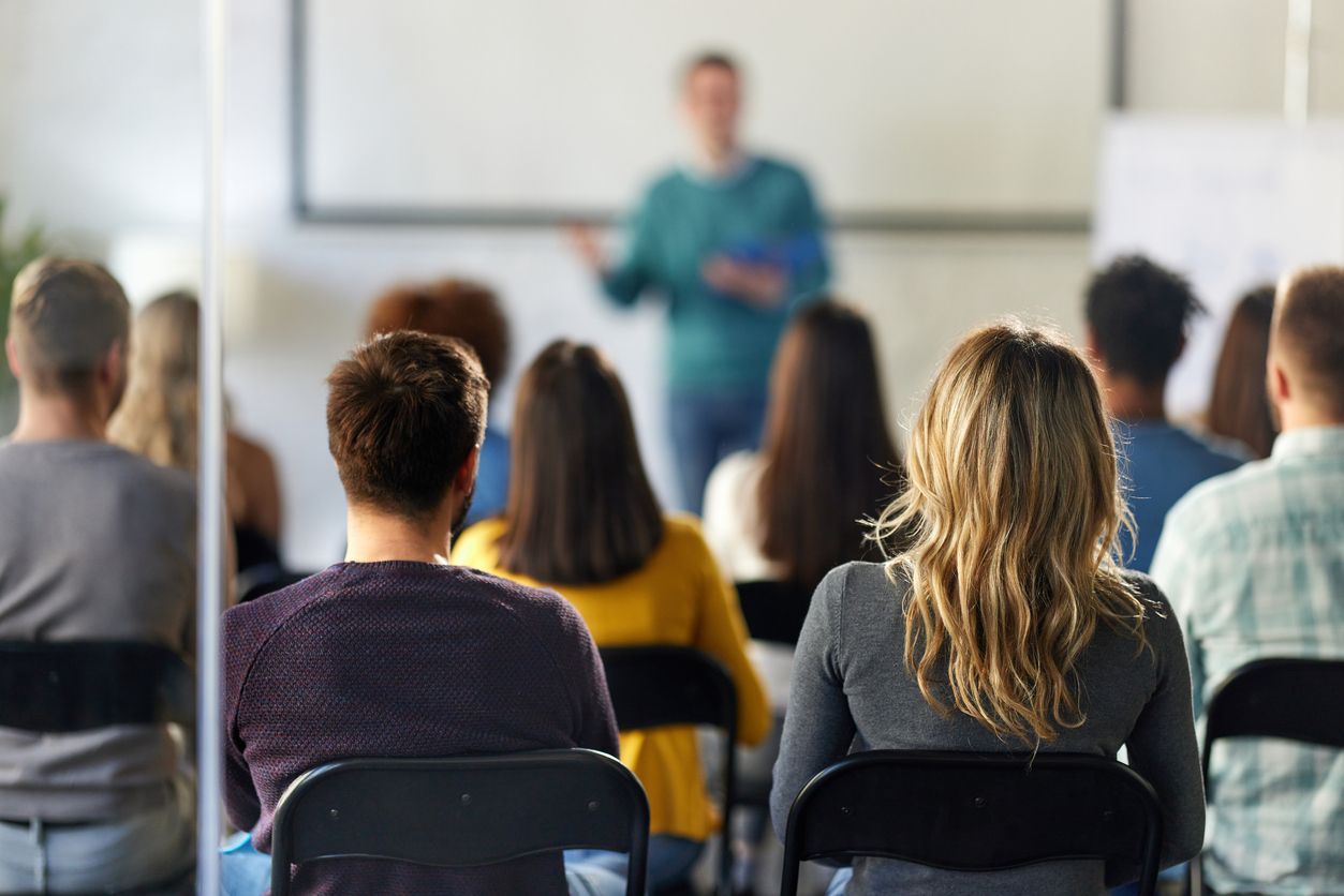 Audience in a classroom facing a speaker, blurred figure in the background, near a screen and white board.