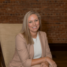 Woman in a tan blazer and white shirt, smiling, seated in a chair, with a brick wall background.