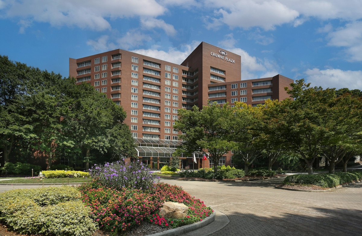 Brown brick high-rise hotel surrounded by trees and landscaping under a blue sky.