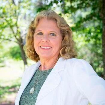Woman with blonde hair wearing a white blazer, smiling in an outdoor setting.