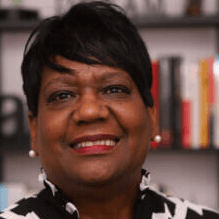 Woman with short dark hair, smiling. Wearing pearl earrings and a black and white patterned top. Bookshelf in the background.