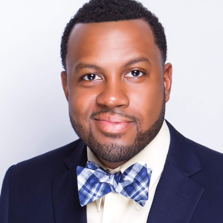 Man in navy suit, plaid bow tie, smiling at the camera, neutral background.