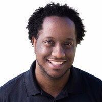 Smiling person with dark skin and curly hair, wearing a black collared shirt, against a white background.