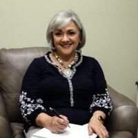 Woman with short gray hair smiles, seated, writing on paper; navy top with floral sleeves.