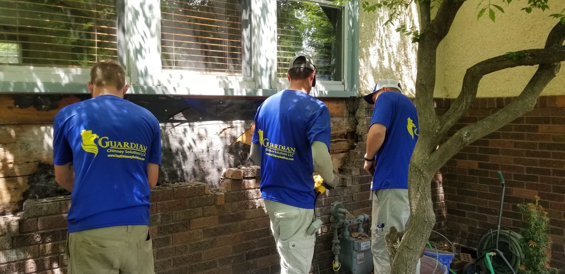 Three men in blue shirts are working on a brick wall.