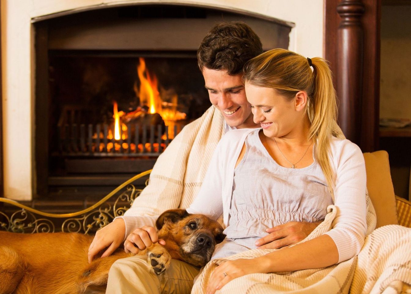 A man and a woman are sitting on a couch with a dog in front of a fireplace.