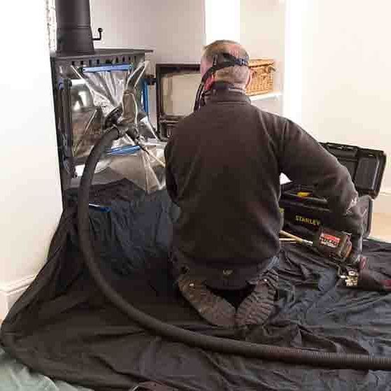 A guardian chimney solutions technician is kneeling down in front of a fireplace with a hose attached to him.