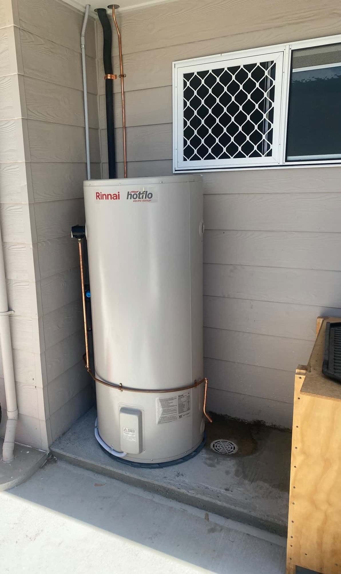 A White Water Heater is Sitting on the Side of a Building Next to a Window — Reed Plumbing Industries in Mackay, QLD