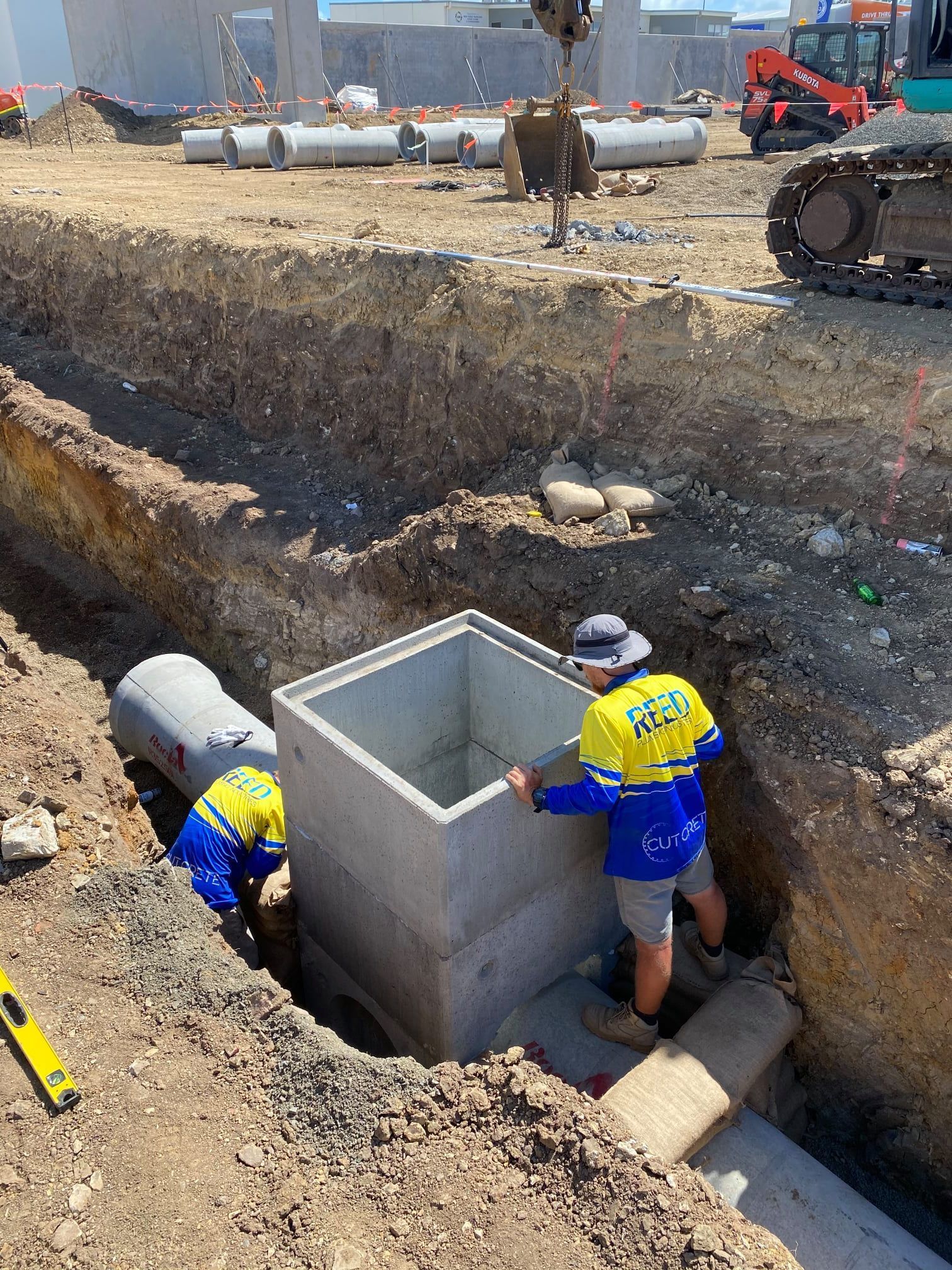 Two  Construction Workers Are Standing in a Trench Looking at a Pipe Being Installed In The Ground — Reed Plumbing Industries in Mackay QLD