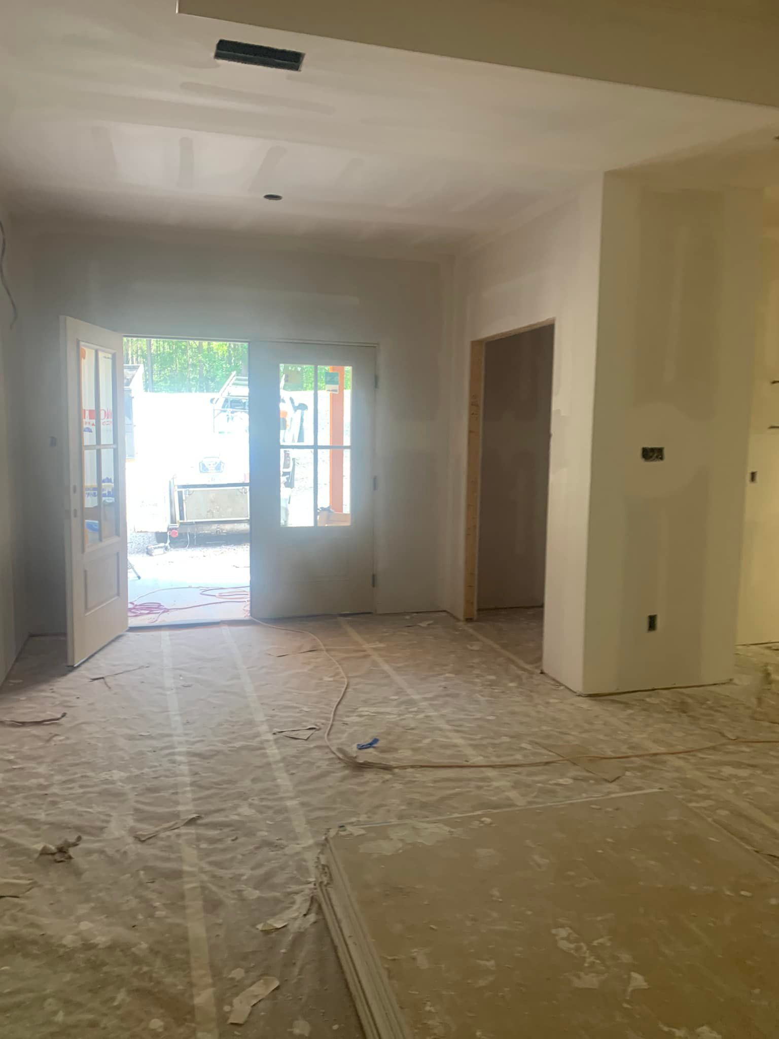 Interior view of a room under construction, with French doors open to an outdoor area and exposed flooring.