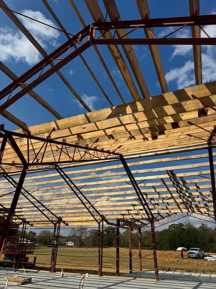 Steel-framed building under construction, with wooden roof supports against a blue sky.