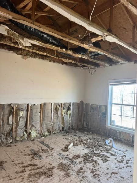 Damaged room: drywall and ceiling missing, exposing beams and insulation. Debris on floor. Window on right.
