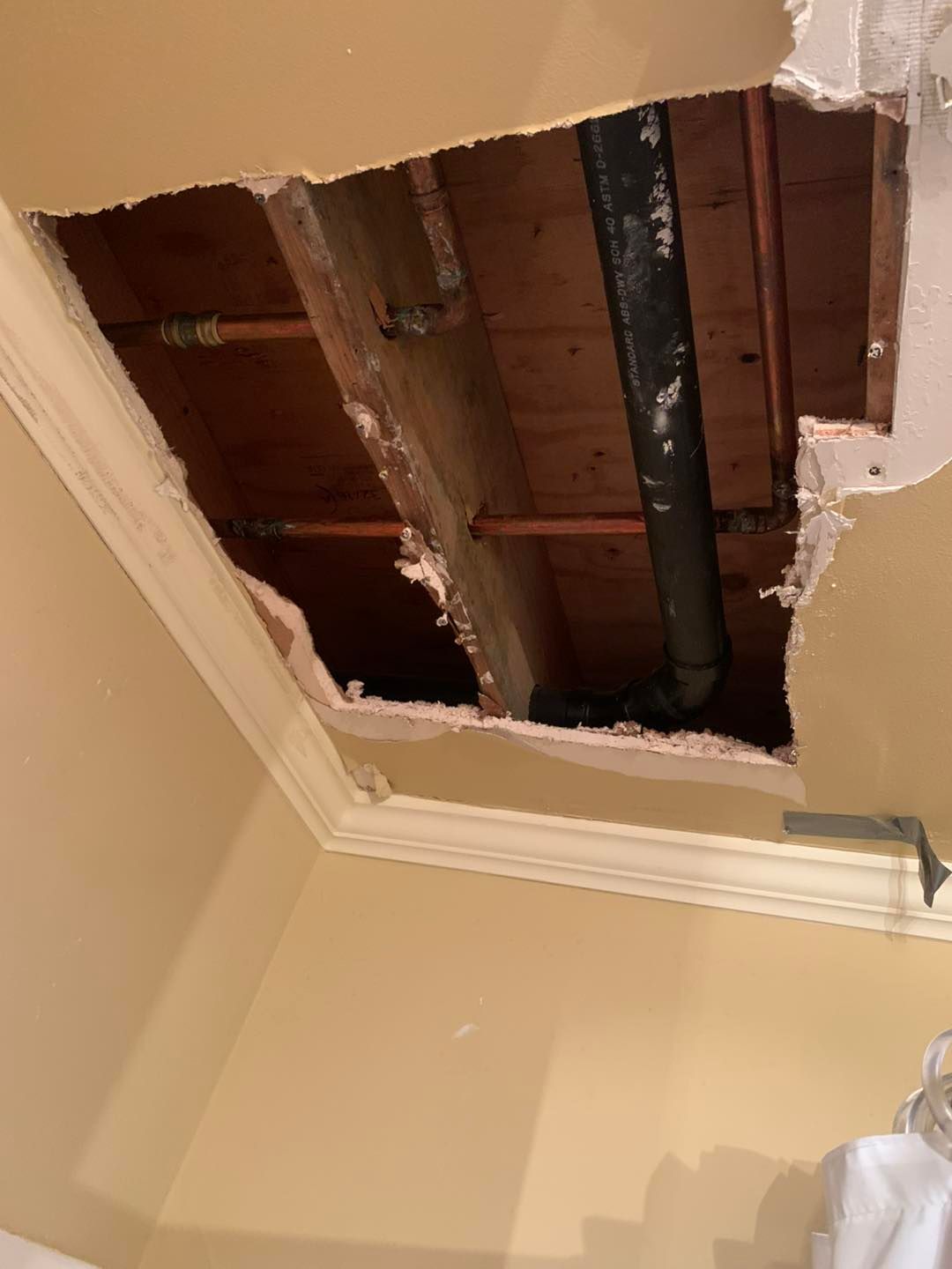 Hole in ceiling reveals plumbing and wood beams. Torn drywall and molding are visible.