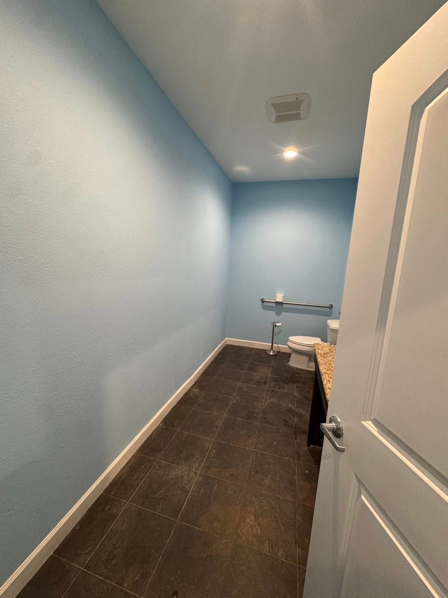 Narrow, blue-walled room with dark floor, open white door in the foreground, and a metal table in the back.