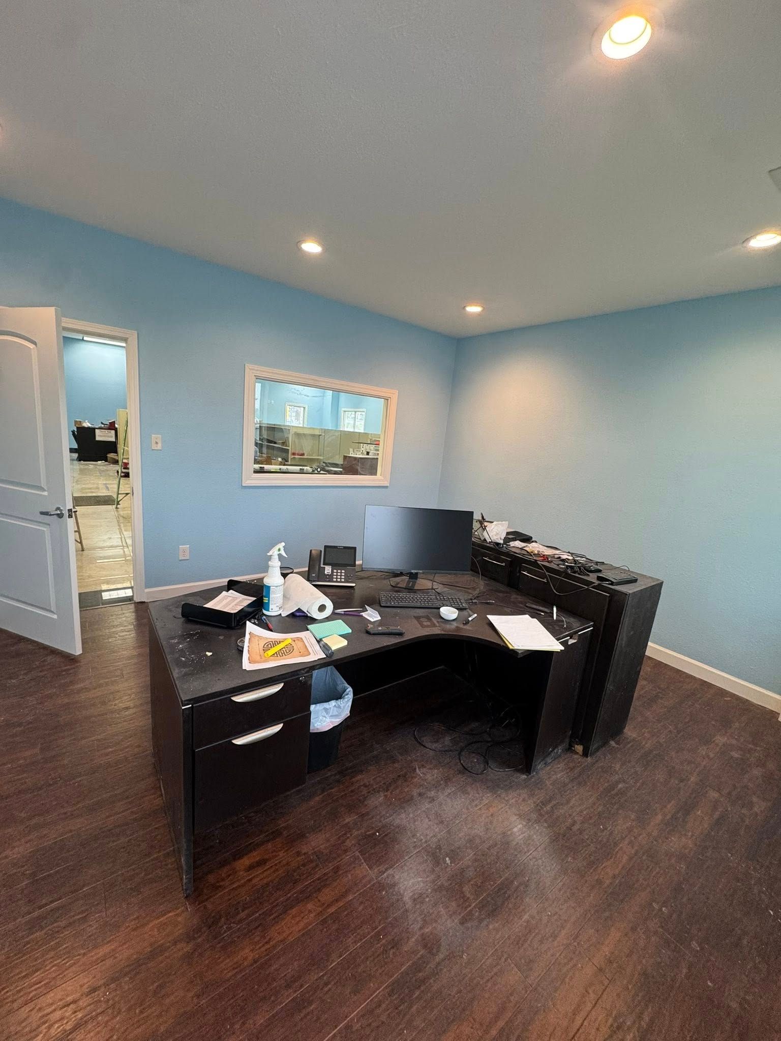 Office with a large black desk, blue walls, a window, and an open door to another room.