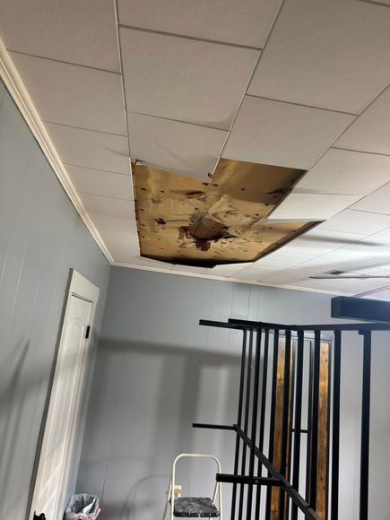 Hole in ceiling with missing tiles, showing damaged wood. Room has gray walls, white trim, and a ladder.