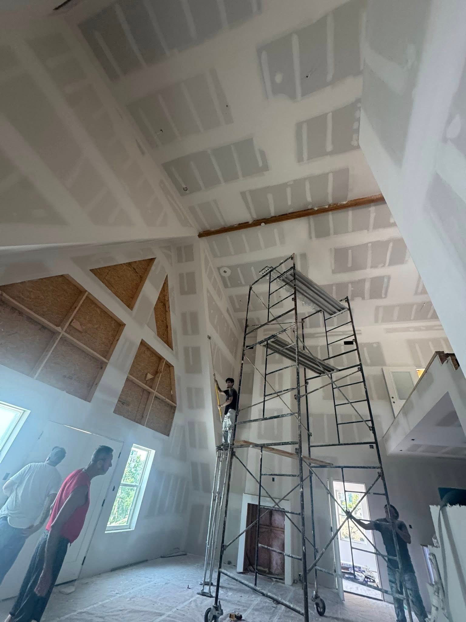 People painting drywall inside a high-ceilinged building. Scaffolding is set up.
