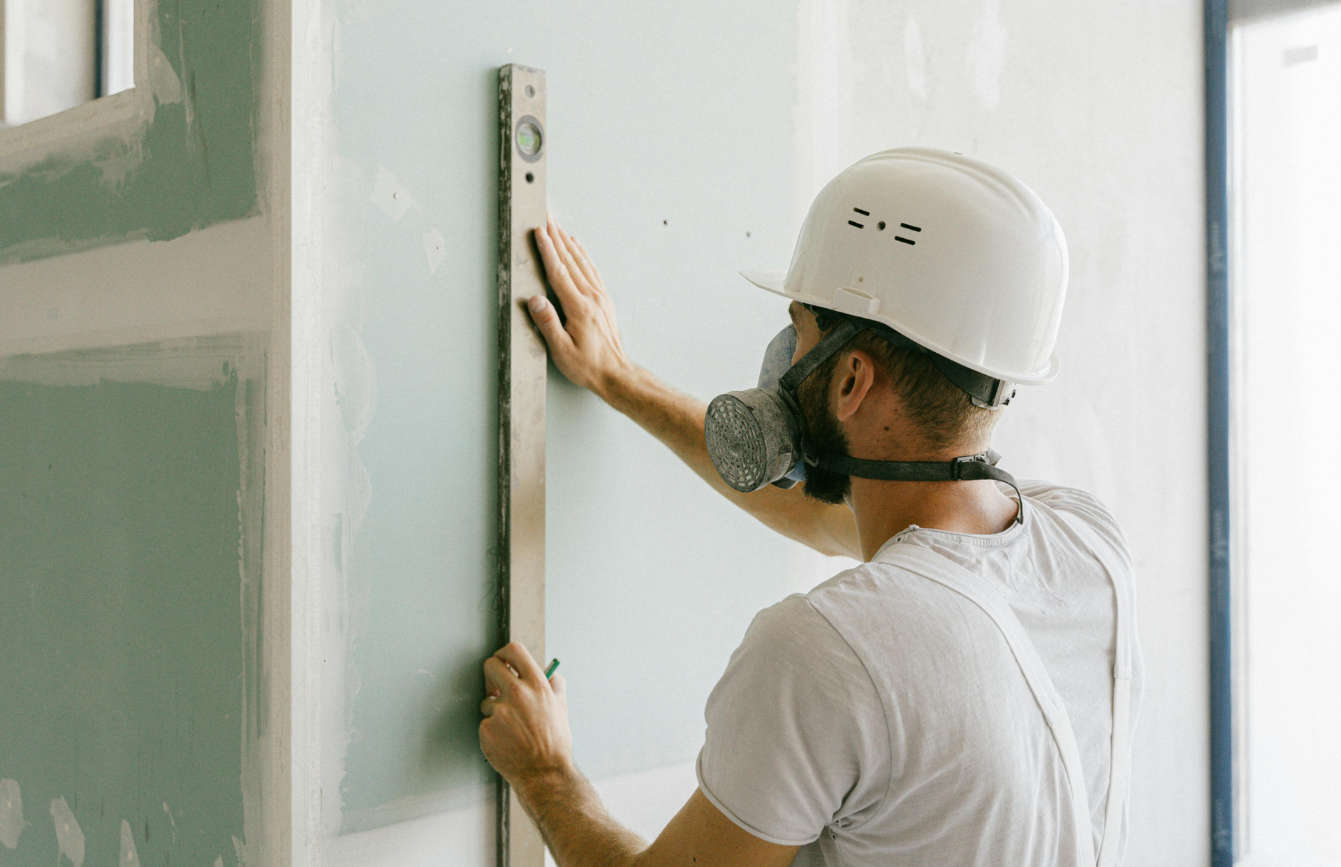 Construction worker in a hard hat and respirator uses a level on a drywall wall.