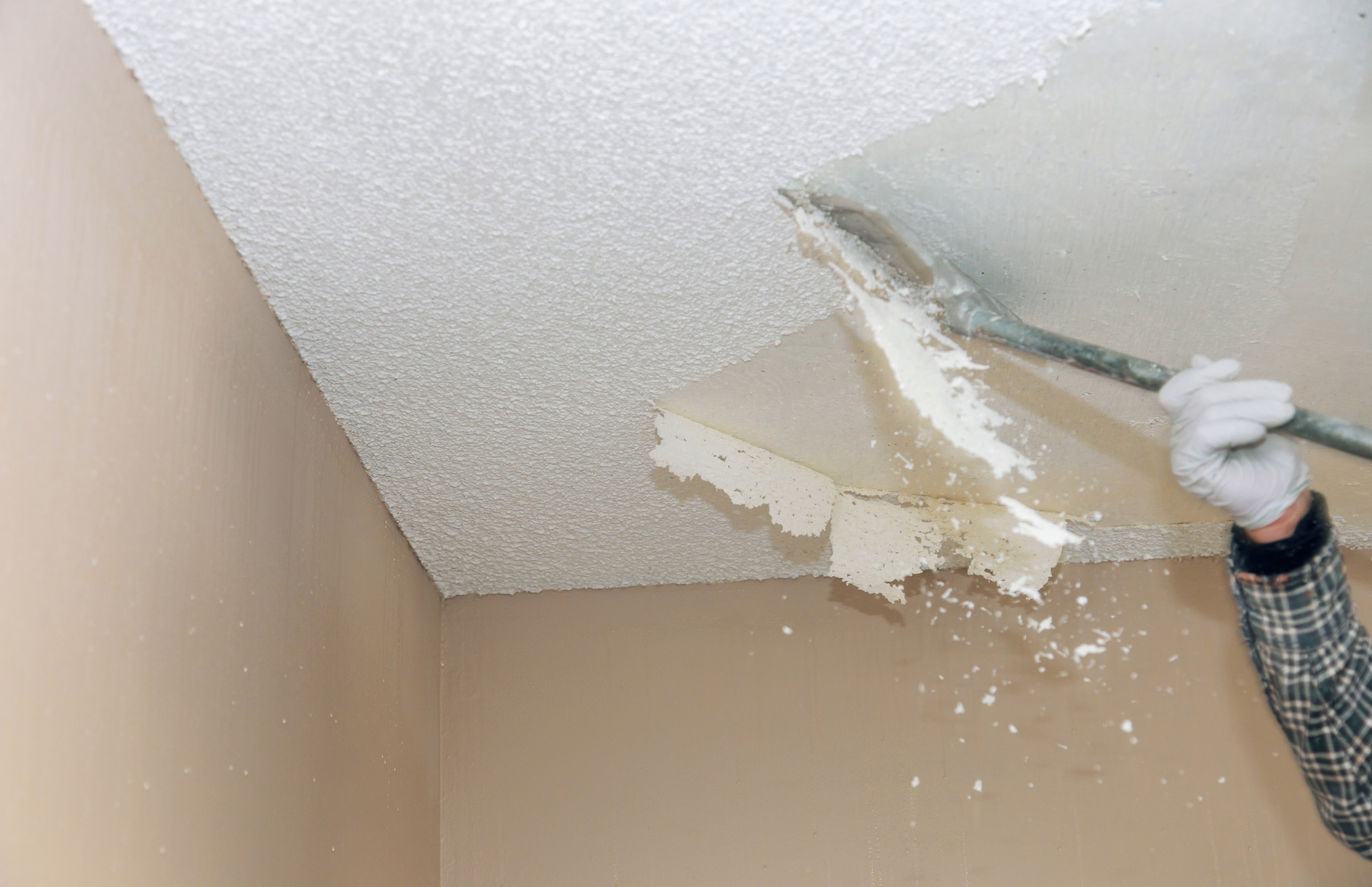 Person scraping textured ceiling with a scraper, removing plaster flakes. Beige walls.