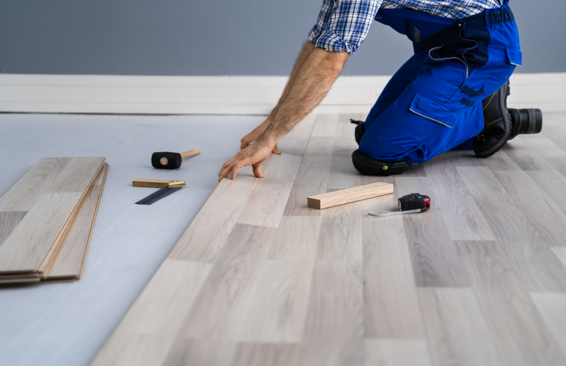Person in blue overalls installing light gray laminate flooring.