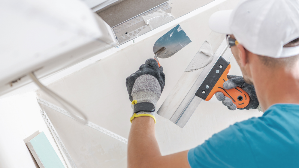 Man in a white cap and gloves using a putty knife to patch drywall on a ceiling.