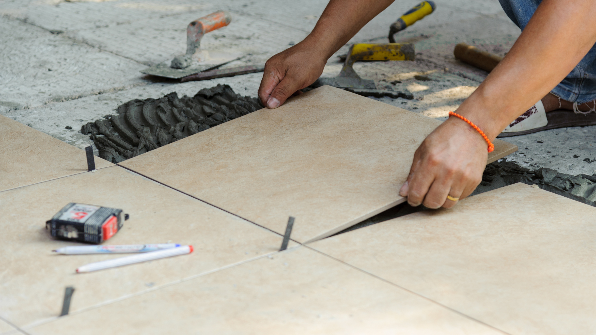Person tiling a floor, using spacers and trowel. Light brown tiles, gray mortar, outdoor setting.