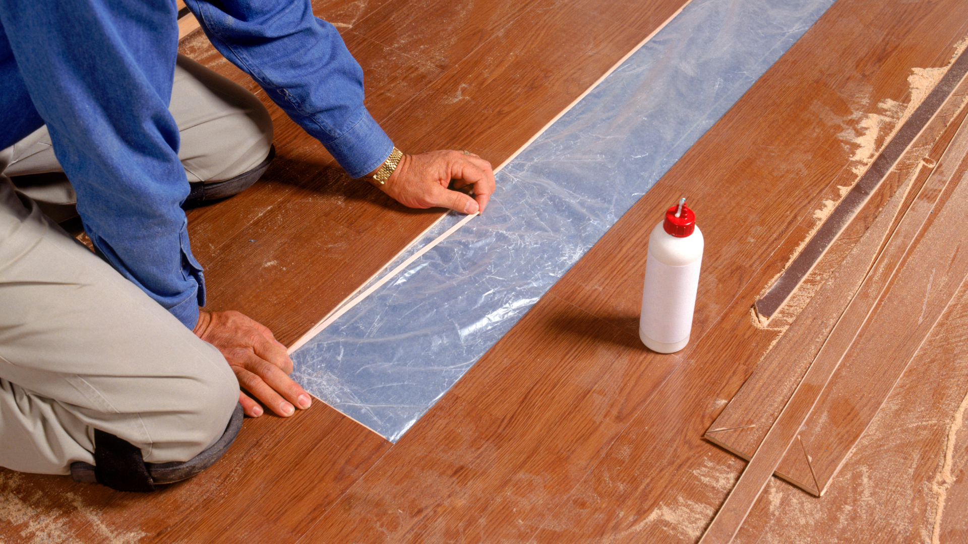 Person kneeling, installing flooring with glue; close-up shows hands, flooring, and bottle of adhesive.