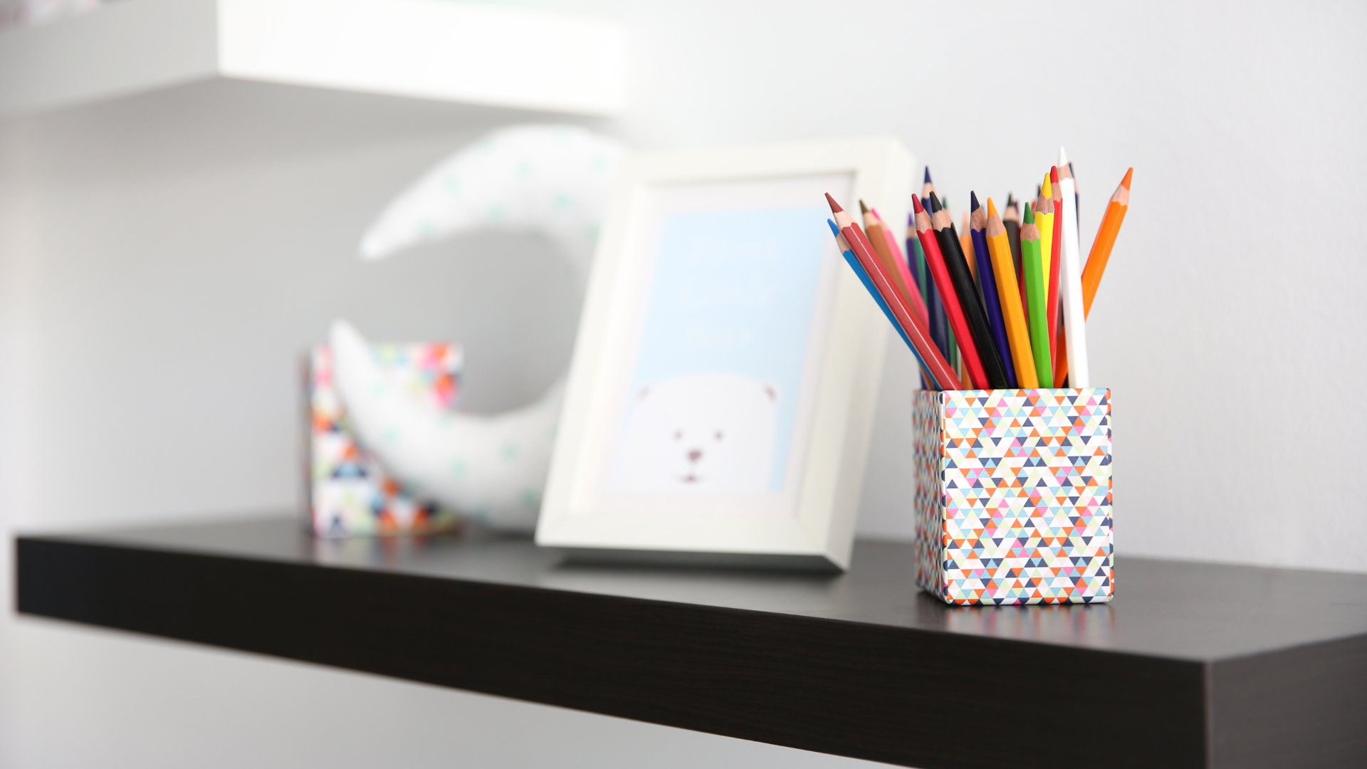 Shelf with art supplies: colored pencils in floral holder, framed picture, crescent moon decoration.