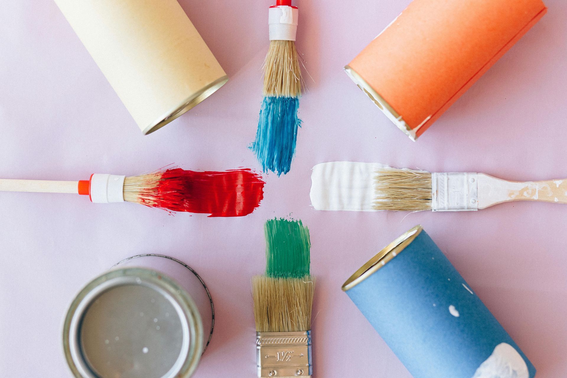 Paintbrushes with blue, red, green, and white paint, surrounded by paint roller covers and a paint can on a pink surface.