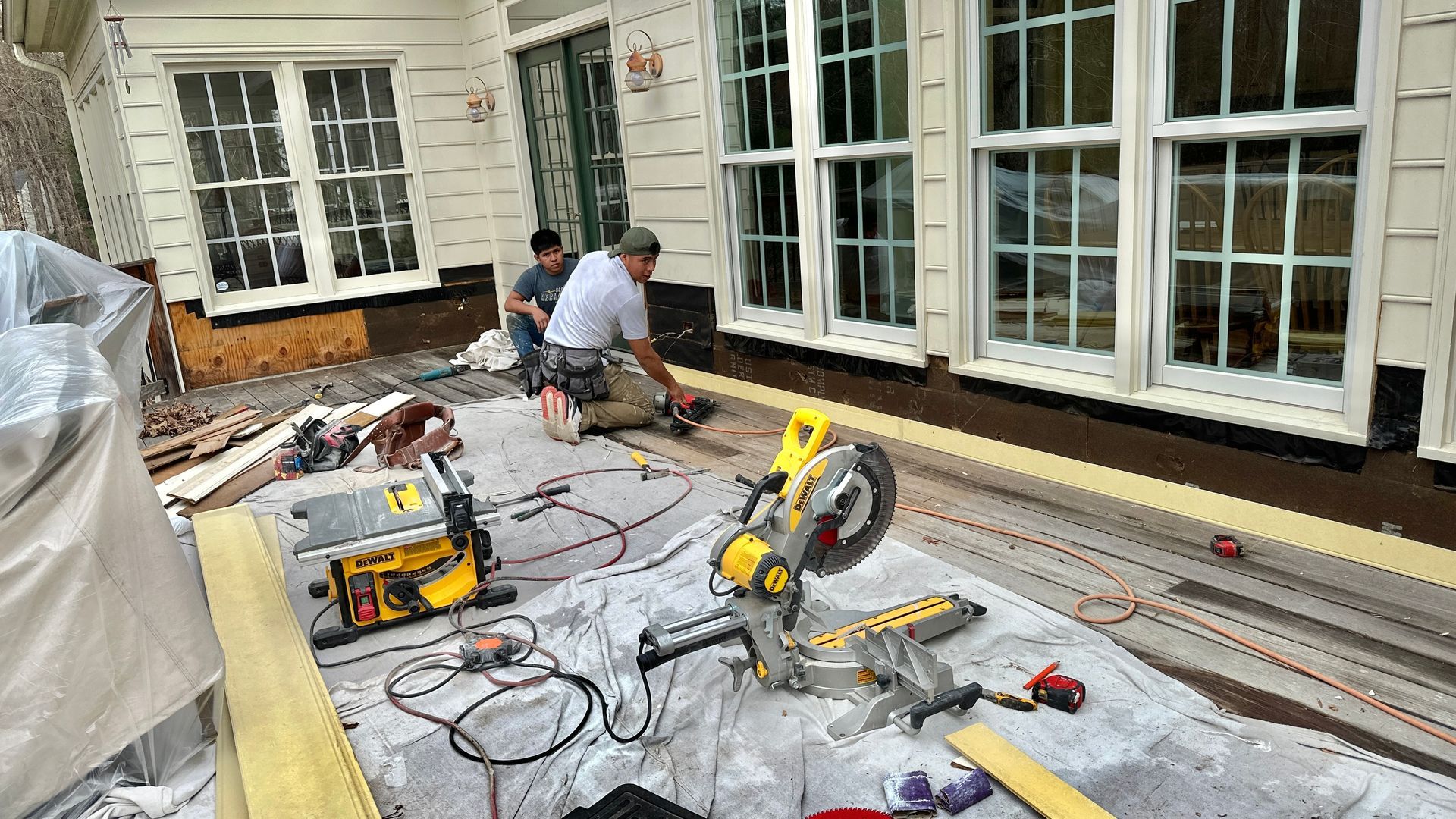 Construction workers using power tools on a deck, near windows. Tools, wood, and tarp visible.